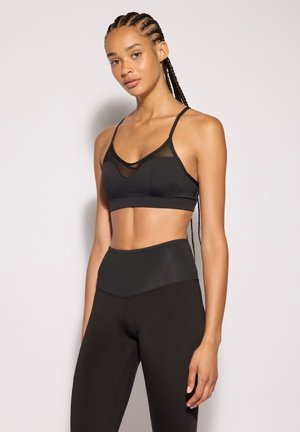 Giovane donna con capelli intrecciati indossa un reggiseno sportivo nero e leggings neri a vita alta, in piedi davanti a uno sfondo chiaro e semplice.