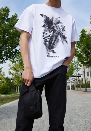 Biała graficzna koszulka z czarnym motywem anioła i napisem "[ Вечность ]" na dole, zestawiona z czarnymi dżinsami i małą czarną torebką.