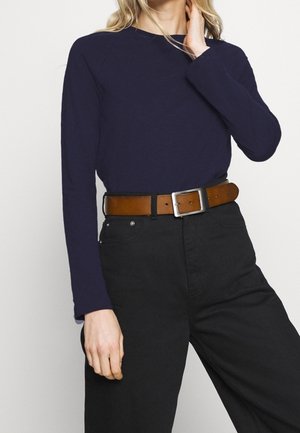 Femme portant une chemise bleu marine à manches longues rentrée dans un pantalon noir taille haute, ceinturée d'une ceinture en cuir marron avec une boucle argentée.