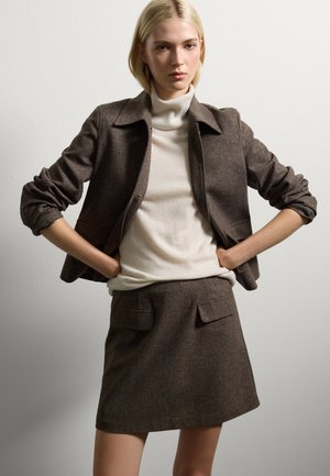 Femme portant une veste marron ajustée et une jupe assortie avec des poches avant sur un pull à col roulé crème, se tenant les mains sur les hanches.