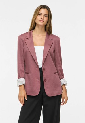 Vrouw staat frontaal, draagt een mauvekleurige blazer met opgerolde mouwen, een witte top en zwarte broek tegen een effen witte achtergrond.