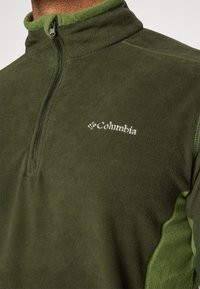 Tamnozeleni flis pulover s polu-zip dizajnom. Ima svjetlije zeleni ovratnik i bočne akcentne detalje, te bijelog Columblinog loga.