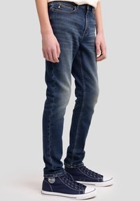 Peenike lõikega tumesinised denim teksad hääldatult kulumisega, millel on klassikaline viie tasku disain ja kontrastsed õmblused. Kombineeritud tumesiniste kanvasist tossudega.