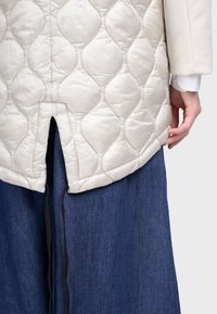 Prešívaná vesta svetlej smotanej farby s okrúhlym spodným lemom a bočným rozparkem, skombinovaná s tmavomodrou džínsovou sukňou. Vesta má hladkú, lesklú textúru.