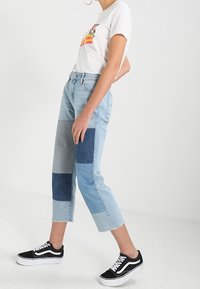 Ljusblå patchworkjeans med varierande nyanser av denim, rå nederkant, kombinerat med en vit t-shirt med tryck och svarta sneakers.