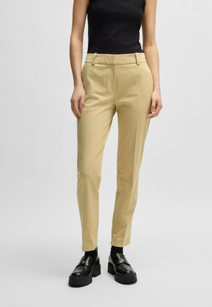 Beige tailored pants with a slim fit, made of smooth fabric. Features a flat front, side pockets, and zippers at the ankle. Worn with black shoes.