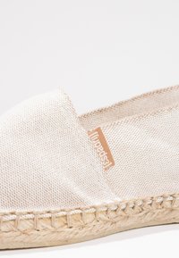Beige canvas espadrille with woven jute sole, featuring a stitched edge and a small brown logo tag. Minimalist design with a smooth texture.
