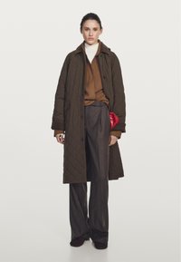 Braun gesteppter Mantel über einem hellbeigen Oberteil, kombiniert mit grauen Weitbein-Hosen. Eine rote Clutch wird unter dem Mantel gehalten. Das Outfit ist geschichtet und hat verschiedene Texturen.
