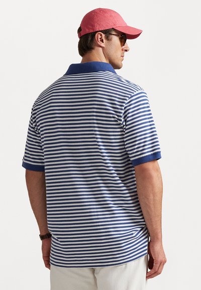 Vīrietis, kurš tērpies zilā un baltā svītrainā polo krekls, bēšu biksēs, sarkanā beisbola cepurē, saulesbrillēs un ar rokas pulksteni, stāv ar muguru pret gaišu fonu.