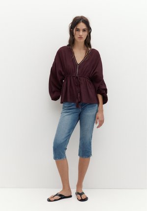 Frau steht vor einer weißen Wand, trägt eine lockere bordeauxrote Bluse, knielange blaue Jeansshorts und schwarze Flip-Flops, mit langen dunklen Haaren offen.