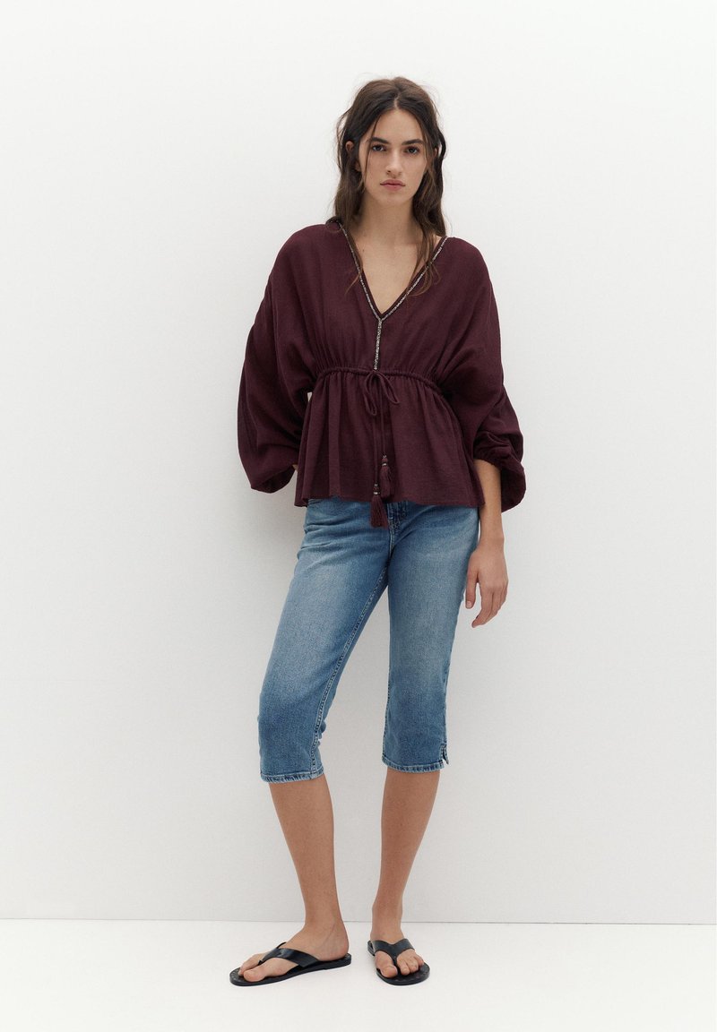 Femme debout contre un mur blanc, portant un chemisier ample couleur bordeaux, un short en jean bleu jusqu'aux genoux et des tongs noires, avec de longs cheveux bruns lâchés.