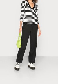 Gestreiftes Longsleeve-Oberteil in Schwarz-Weiß mit Puffärmeln, kombiniert mit weiten schwarzen Hosen und weißen Plateau-Stiefeln, hält eine kleine grüne Tasche.