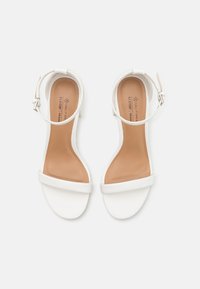 Sandales à talons en cuir vegan blanc avec une finition lisse, bout ouvert, large bande à l'avant et fermeture à boucle au niveau de la cheville. Semelle intérieure beige avec marque.