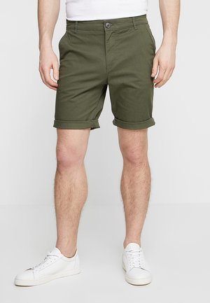 Homme portant un short vert olive avec ourlet retroussé, un t-shirt blanc et des baskets blanches, debout devant un fond blanc uni.