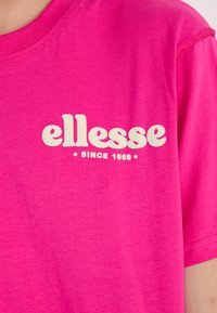 T-shirt rose vif à manches courtes avec un grand logo blanc "ellesse" et le texte "SINCE 1969" en format courbé. Tissu doux.