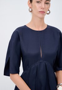 Laivastonsininen mekko, jossa on pyöreä pääntie, avoin etuleikkaus ja lyhyet hihat. Mekossa on poimuja ja sileä pinta, ja se on yhdistetty suuriin hopeisiin renkaisiin korvakoruihin.