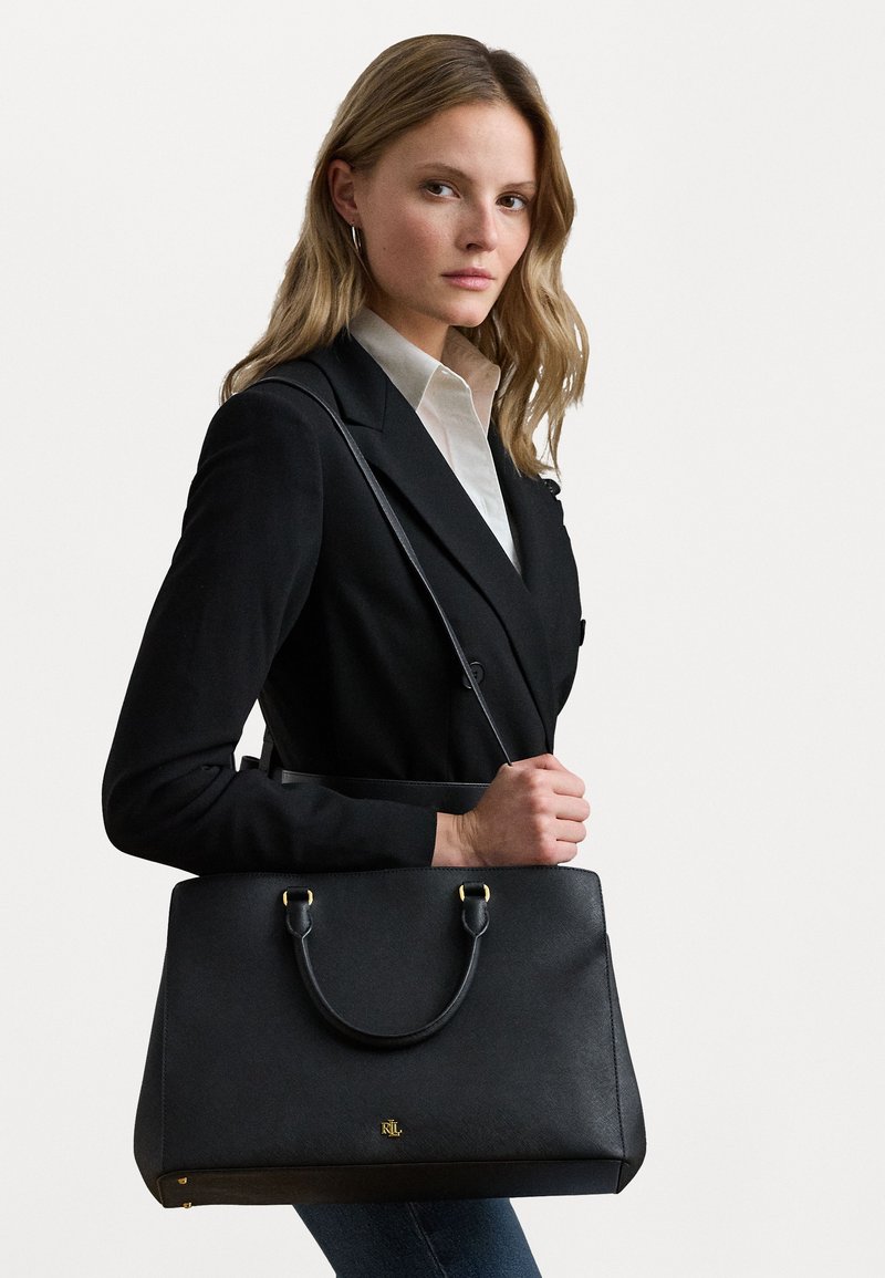 Ženska v črnem blazerju in beli srajci nosi veliko črno usnjeno torbo z zlatim logotipom in dvema ročajema čez ramo.