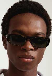 Lunettes de soleil avec une monture en écaille épaisse et des verres sombres. Texture de surface lisse avec un design angulaire, reposant sur le visage d'une personne.