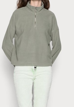Femme portant un pull polaire à demi-fermeture éclair vert clair et un pantalon blanc avec des détails subtils de teinture nouée verte, debout devant un fond uni.