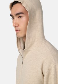 Sweat à zip beige avec une texture douce, présentant un col montant et une capuche. Le design est d'un style minimaliste avec des détails de couture subtils.