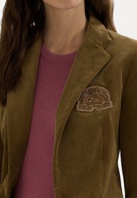 Žena oblečená v olivově zeleném kordovém saku s vyšitým emblémem přes tlumený fialový pletený svetr.