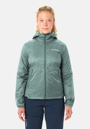 Junge Person mit lockigem Haar trägt eine helltürkisfarbene Kapuzenjacke, die hochgeschlossen ist, und navyblaue Hose, steht vor einem einfarbigen Hintergrund.