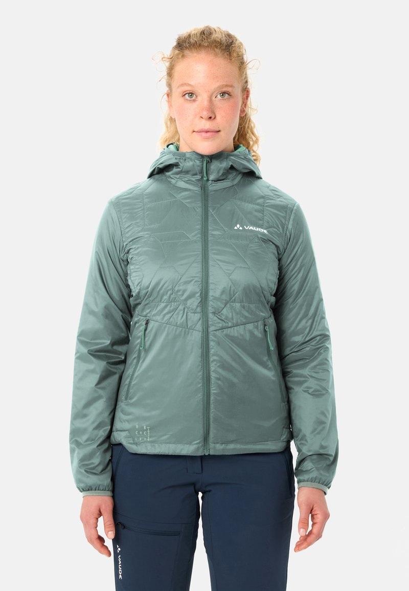 Junge Person mit lockigem Haar trägt eine helltürkisfarbene Kapuzenjacke, die hochgeschlossen ist, und navyblaue Hose, steht vor einem einfarbigen Hintergrund.