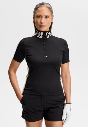 Vrouw die een zwarte golfoutfit draagt met een witte kraag met patroon, zwarte korte broek en een witte golfhandschoen aan de rechterhand, staand met handen in de zakken.