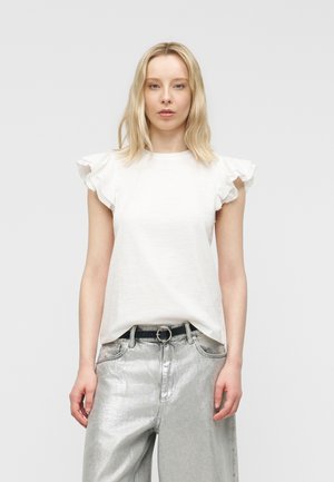 Svetlolaska ženska, oblečena v bel zgornji del z nabranimi rokavi, zataknjen v visoke kovinske srebrne hlače s črnim pasom, stoji na enostavnem ozadju.