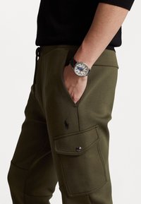 Olive green jogger pants with a side pocket, featuring a dark logo, and a silver watch with a multicolored strap on a hand.
