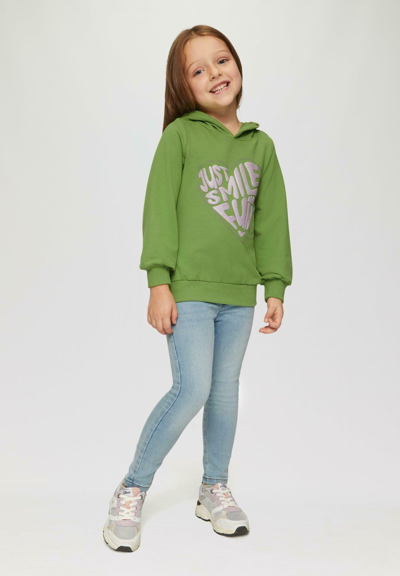 Groene hoodie met een hartgrafiek waarop "Just Smile" in zilver staat, gecombineerd met lichtblauwe skinny jeans en grijze sportschoenen.