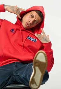 Jeune homme avec des tatouages portant un sweat à capuche rouge avec le texte "Authentic", qui pointe la capuche tout en levant une jambe vers l’avant montrant la semelle de la chaussure.