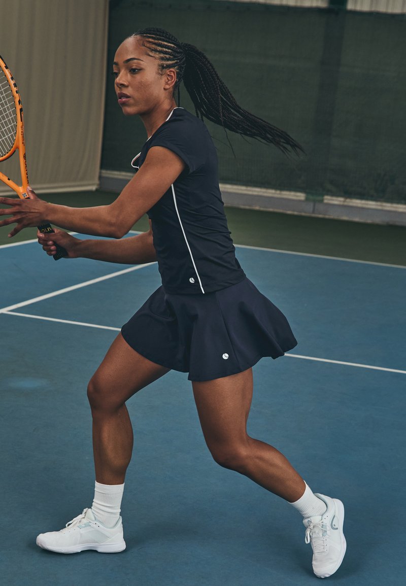 Tennis outfit with a fitted navy top and flared navy skirt. The player wears white athletic shoes and white ankle socks, holding an orange racket.