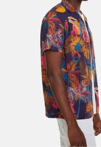 Camicia a maniche corte di colore blu navy con una vivace stampa tropicale che presenta foglie di palma e uccelli. Design con bottoni e vestibilità comoda.