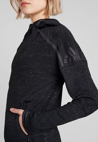 Czarna kurtka zapinana na zamek z teksturowanej tkaniny, dopasowany krój, kaptur i logo Adidas na ramieniu. Posiada kieszenie boczne i elastyczne mankiety.