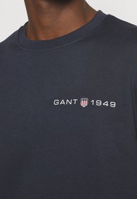 Gros plan d’une chemise en coton bleu marine foncé avec « GANT 1949 » et un petit logo en forme de bouclier rouge, blanc et bleu sur la poitrine.