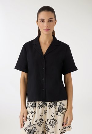 Donna con capelli castani legati in una coda di cavallo, che indossa una camicia nera a maniche corte con bottoni e pantaloncini crema con motivo floreale nero.