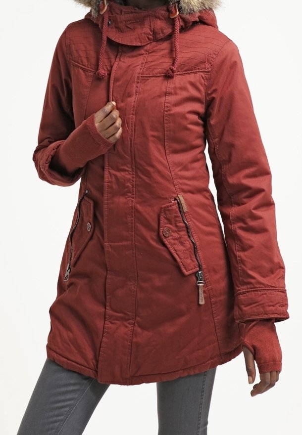 Personne portant un manteau d’hiver couleur rouille avec une capuche en fourrure, des poches zippées et des poignets avec trous pour le pouce, associé à un pantalon gris.