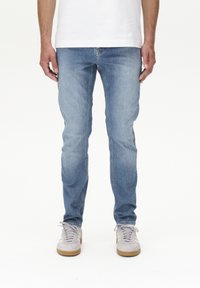 Világoskék, slim fit farmernadrág, klasszikus öt zsebes dizájnnal és finom kopással. Szürke sportcipővel párosítva.