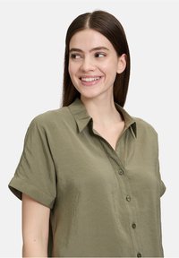 Olijfgroene shirt met korte mouwen, knoopsluiting, kraag en een lichte, gekreukte textuur. De stof heeft een subtiele glans.