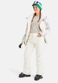 Skiausrüstung mit einer weißen Jacke mit floralen Akzenten, einem beigen Oberteil, cremefarbenen Hosen und schwarzen Skischuhen. Zubehör umfasst eine grüne Mütze und eine Skibrille.