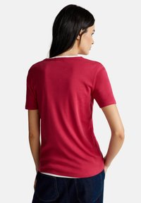 Donna con capelli lunghi e scuri, che indossa una maglietta rossa a maniche corte e jeans blu scuro, vista di spalle su uno sfondo bianco.