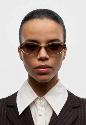 Mujer con el cabello peinado hacia atrás, que lleva gafas de sol ovaladas marrones, camisa blanca con cuello y chaqueta de traje oscura a rayas, sobre un fondo liso.