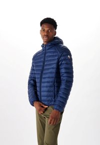 Chaqueta acolchada azul marino con cierre frontal y capucha, que presenta una sutil textura acolchada y un detalle del logo en la manga. Combinada con pantalones verdes.