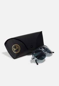 Ochelari de soare Ray-Ban cu lentile gri într-un cadru transparent, însoțiți de o carcasă din piele neagră texturată cu un logo circular auriu.