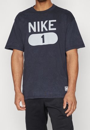 Mężczyzna noszący czarną koszulkę Nike z dużym białym napisem "NIKE 1" na przodzie oraz małą naszywką z logo w pobliżu dolnego brzegu.