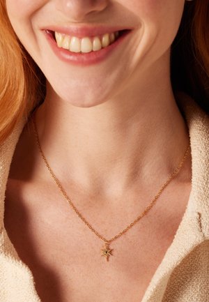 Collier en or avec un pendentif en forme d'étoile, orné de petites pierres précieuses. Chaîne délicate avec des maillons, portée sur un teint clair.