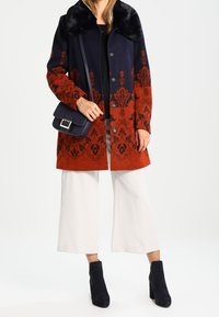 Uldblandingsfrakke med en sort pelskrave, prydet med et blåt og rødlige-orange blomsterdesign. Saten med hvide vide bukser og sorte ankelstøvler.