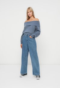 Ανοιχτό μπλε ανδροκούμπο πουλόβερ με ριμπά υφή, συνδυασμένο με φαρδιά τζιν παντελόνια σε μπλε απόχρωση και μαύρα αθλητικά παπούτσια. Απλή, casual εμφάνιση.