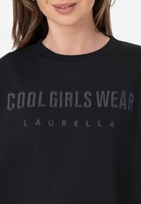 Sudadera negra con el texto "LAS CHICAS COOL LUCEN LAURELLA" en una impresión tonal. Cuello de canalé y textura de tejido suave.